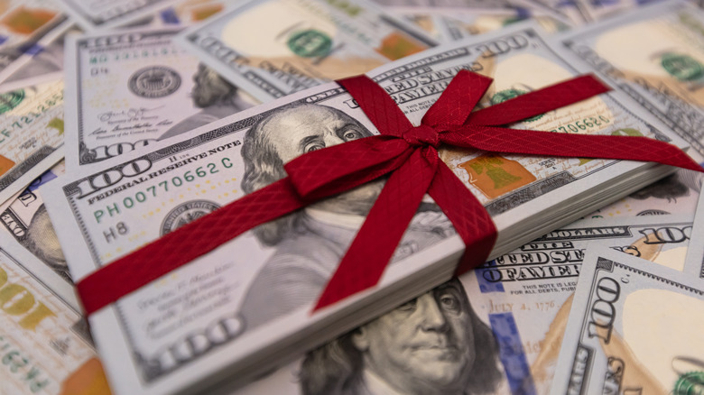 A stack of 100 dollar bills with a red ribbon tied around it and loose bills spread out across the background.