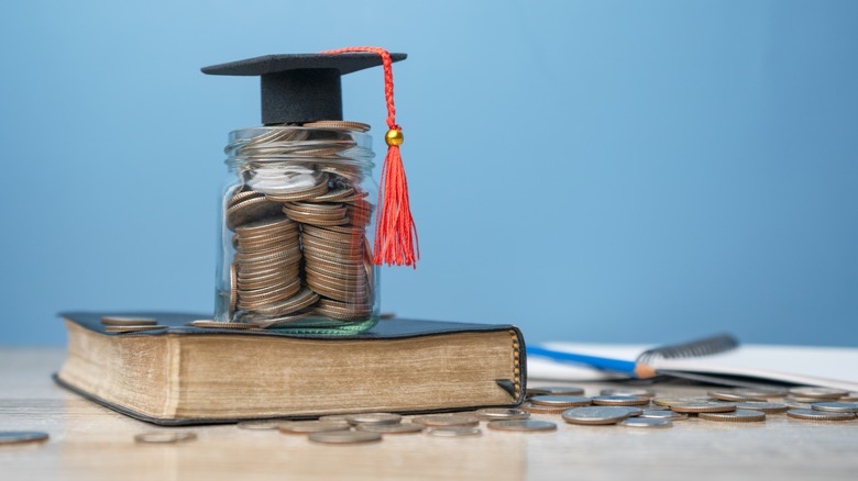 A closed book sitting on a table surrounded by a notebook and loose change, and a glass jar of coins with a small graduation cap sitting on top of it resting on top of the book.