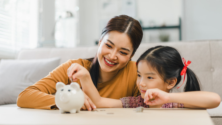 A smiling mother and daughter placing a coin into a pink piggy bank.