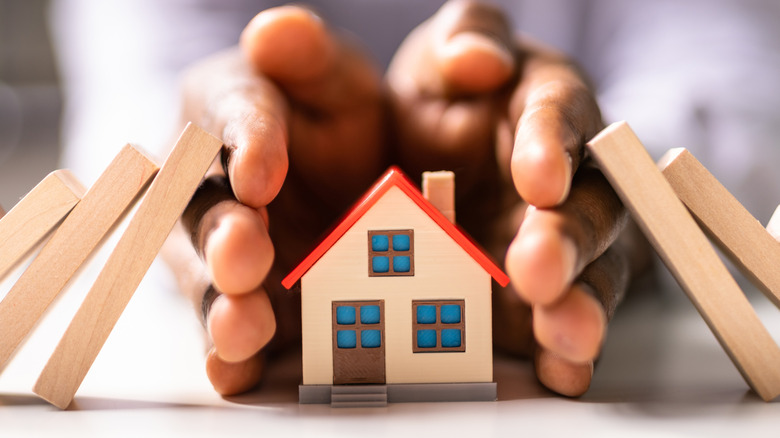 Hands protecting a miniature home from falling dominos.