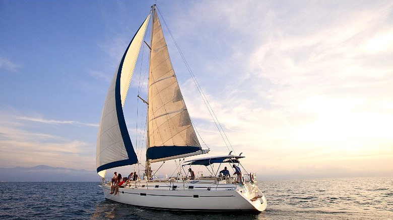 A white sailboat with white sails with blue trim on the open water with some people sitting on the edge of the boat and other people standing on the boat