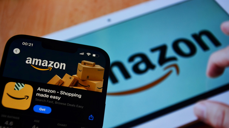 A closeup shot of a cellular phone with the Amazon shopping app displayed on the menu and a larger tablet in the background showing the Amazon logo.