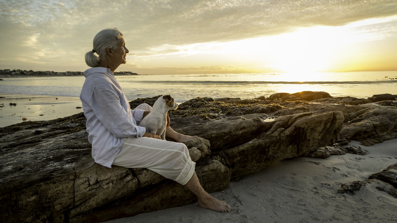 An elderly woman sitting on a rock watching the sunset at a beach.