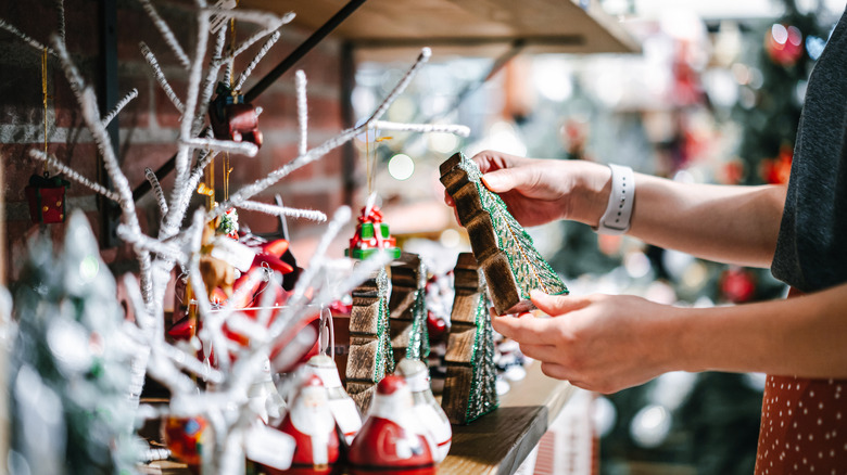 A pair of hands hold a Christmas tree decoration, just plucked from a festive display of holiday decor in a brightly-lit store aisle.