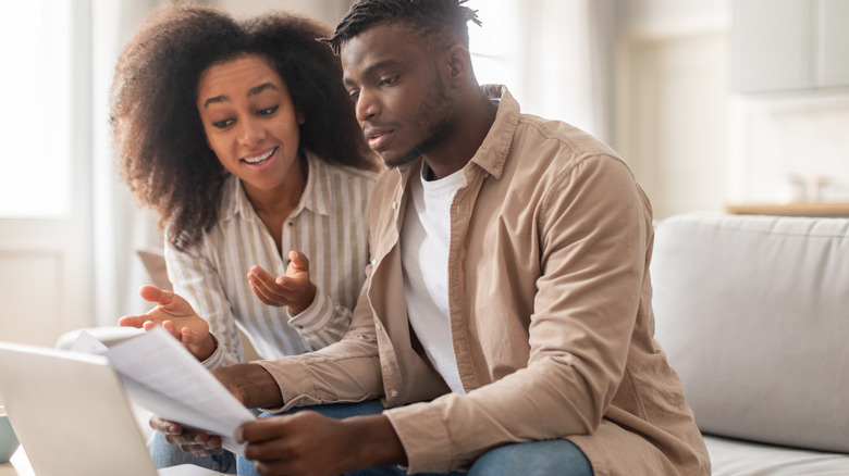 A man and woman sitting on a couch holding documents in front of an open laptop.