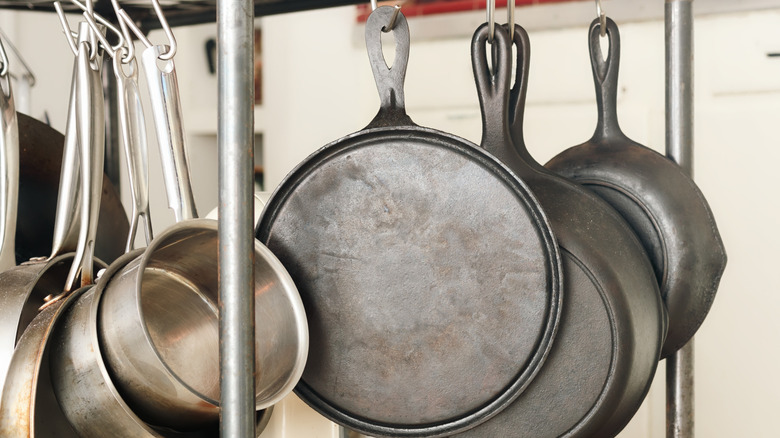 Pots and cast iron skillet pans hang from a rack, against a white background.