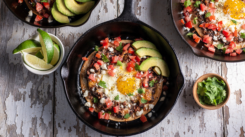 A colorful display of huevos rancheros in cast iron skillets, served on an old wooden picnic table.