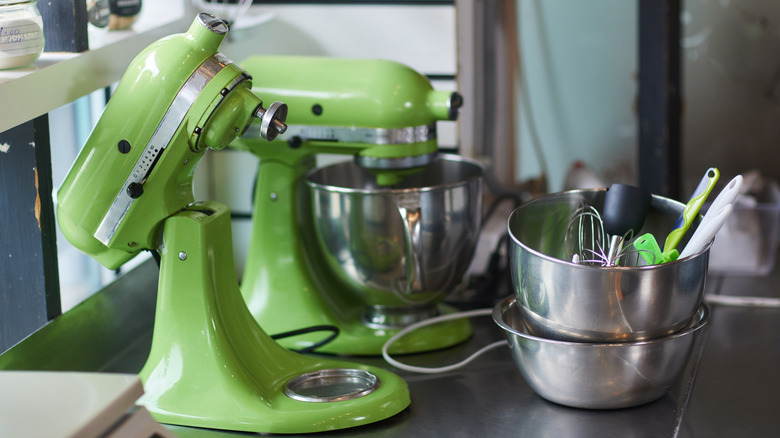 Two green KitchenAid mixers sitting next to a bowl of attachments.