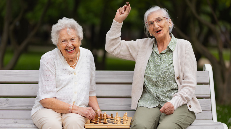 Two smiling elderly woman playing chess on a park bench