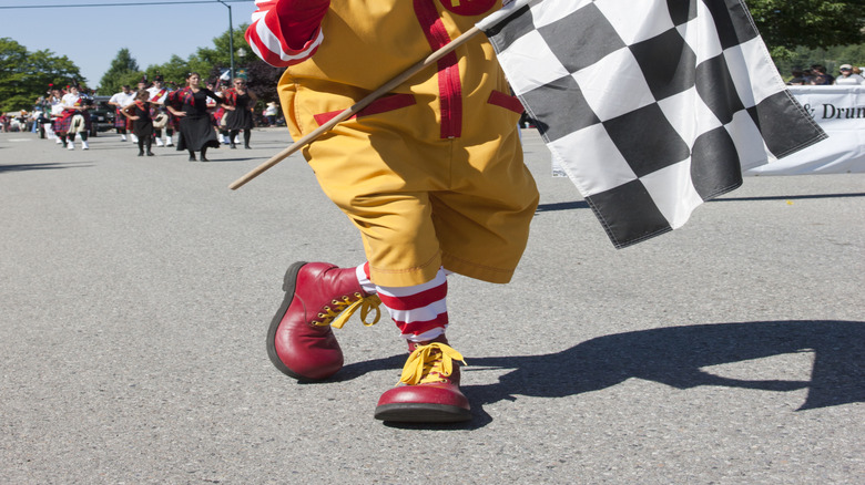 The iconic Ronald McDonald flashes a gloved thumbs up with one hand and holds a racing flag in the other at an organized run/walk event outside.