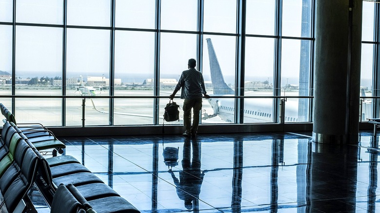 Man holding a backpack in left hand looks out airport window at planes