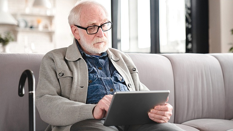 Older person sits on a couch using a tablet device with a cane next to him