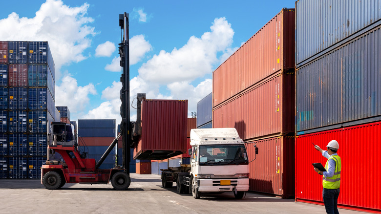 A man in a yellow caution vest oversees the loading of a shipping container onto a drayage vehicle, in a busy shipyard on a blue-sky day.