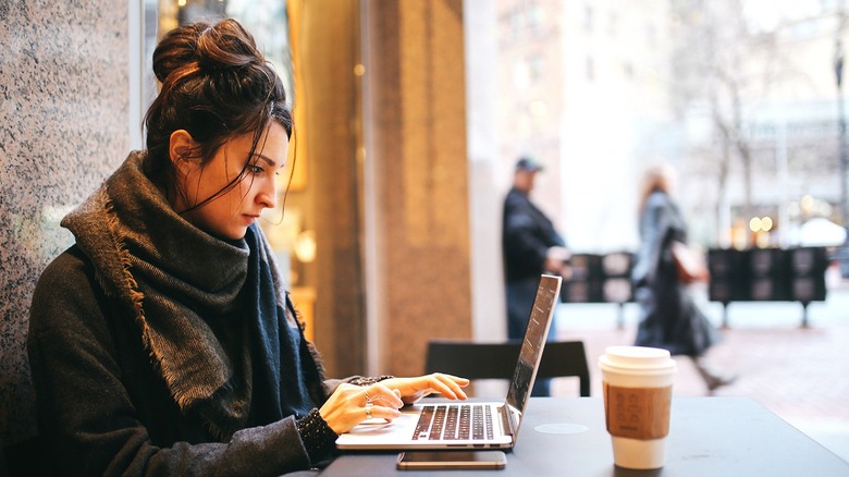 Person wearing a scarf and coat types on laptop at a table outside with a coffee and phone nearby