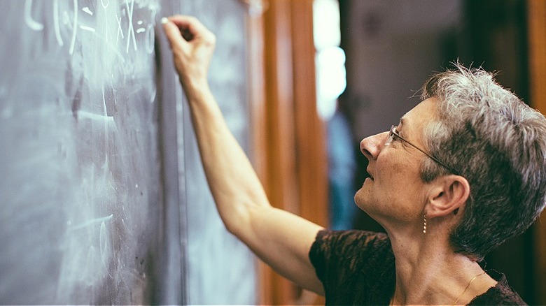 An older female math teacher smiles as she writes an equation on a dusty chalkboard