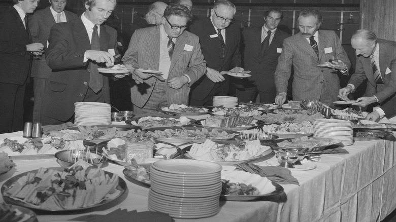 Younger men watch as old men take food from buffet