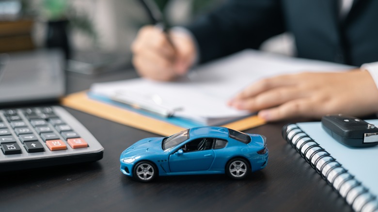 A toy car on a desk with a calculator and someone working in the background.