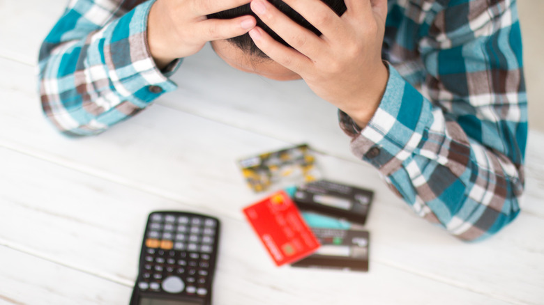 A man holding his head in his hands while he looks at credit cards and a calculator.