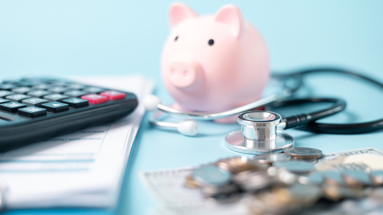 A pink piggy bank, a calculator, and a stethoscope surrounded by blurry images of coins, paper currency, and a stack of papers.