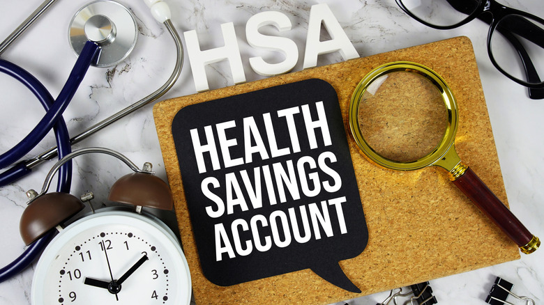 A rectangular cork board with a magnifying glass and a thought bubble with the words 'Health Savings Account," surrounded by an alarm clock, a stethoscope, a pair of glasses, and the letters "HSA."