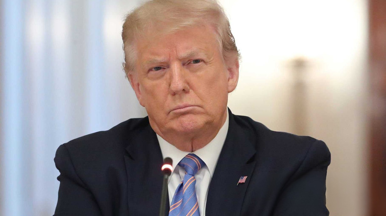 Donald Trump frowns with arms crossed, wearing an American flag pin, in front of a bright backround.