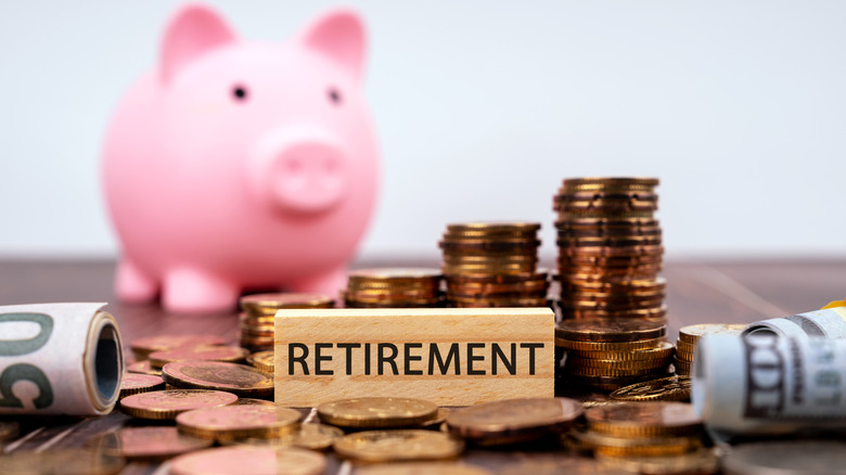 A small wooden sign with the word "Retirement" printed on it surrounded by stacks of coins and rolled up bills with and the blurred image of a pink piggy bank in the background.