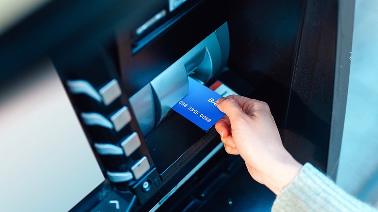 A person wearing a tan sweater placing a blue credit card into a black and silver ATM