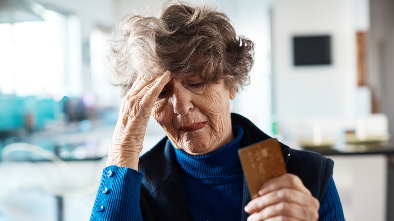 A woman with gray hair holding a credit card in one hand and resting her forehead on the other hand in a sign of regret.